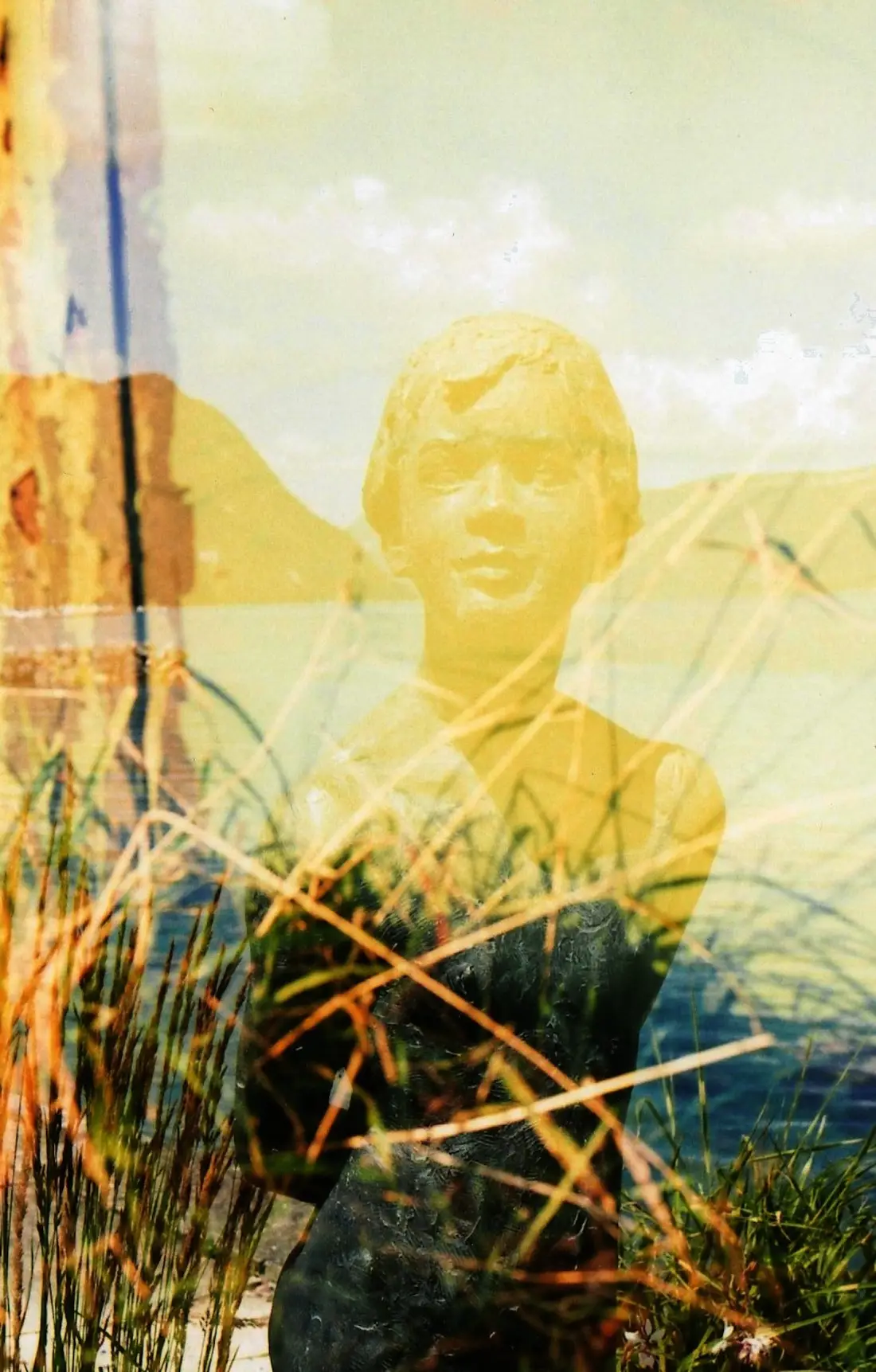 Photographie en surimpression. On y voit des herbes rousses superposées à une statue, ou une sculpture, au bord de la mer près d'un rocher.